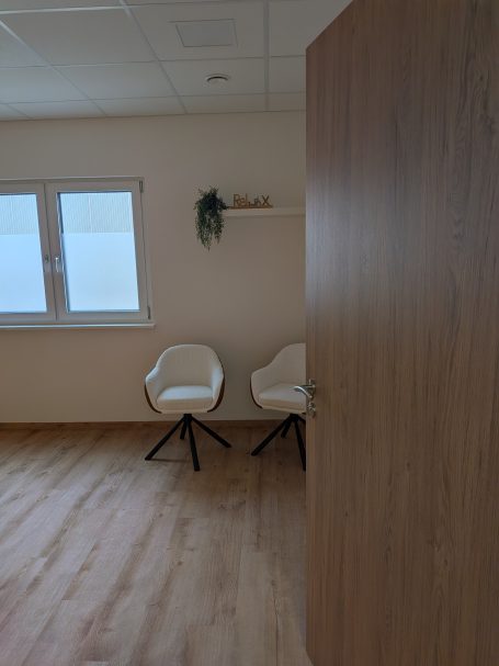 Heller Raum mit Holzfußboden, zwei weißen Stühlen und einem Regal an der Wand.